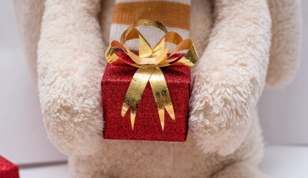 A cozy teddy bear holding a red gift box with a gold ribbon.
